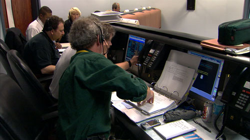 In this image, six individuals sit at consoles in front of computers during a launch rehearsal for the Mars Reconnaissance Orbiter mission.  Launch vehicle manager Arden Acord (a gray-haired, Caucasian middle-aged man with a goatee) points to data on project manager Jim Graf's computer.  Graf is a brown-haired Caucasian man in his fifties.  Flight system manager Howard Eisen sits to Graf's left.  He is a Caucasian man with brown hair and a goatee.  Next to Eisen are two Lockheed Martin employees, one of whom is Tammy Harrington, the mission integration manager.  She is a blond woman in her forties.