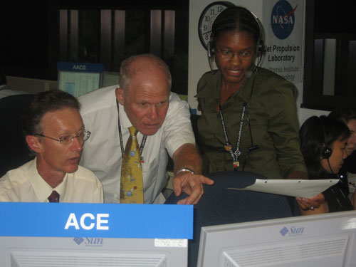 This photo shows team members hover in front of the computer screens in the mission support area.  Two men wearing ties and one young African American woman wearing a headset (to talk to mission control) point at the screens and compare notes from printed out spacecraft predicts made before launch.  Two women in the far right of the image talk earnestly about other data coming down from the spacecraft.