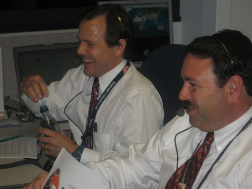 This photo shows Todd Bayer and Dan Johnston, both wearing white shirts, ties, and headsets, laugh and smile over the successful launch.  Todd is putting the cap on a water bottle, and Dan is flipping through a flight plan.