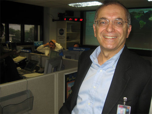 This photo shows Dr. Charles Elachi smile in front two 3-foot computer screens displaying the data and Earth Ground-Track Map relating to the MRO launch.  Dr. Elachi has glasses, salt-and-pepper hair, and wears a blue shirt with a dark blue jacket.  Dan Johnston is in the background at a computer, putting on a headset.