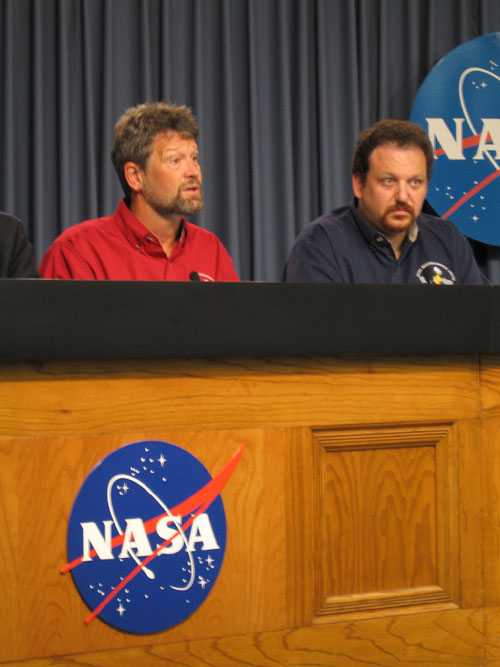In this image, Jim Graf (Mars Reconnaissance Orbiter project manager) is seated on the left at a long, wooden table bearing the circular red, white and blue NASA 'meatball' logo.  He is wearing a long-sleeved red, button-up shirt and answering a question from a reporter.  He is a middle-aged Caucasian man with brown hair, a goatee and a light beard.  On the right is flight system manager Howard Eisen.  He is a Caucasian man in his thirties or early forties with brown hair and a goatee. He is wearing a dark blue, button-up, long-sleeved shirt.