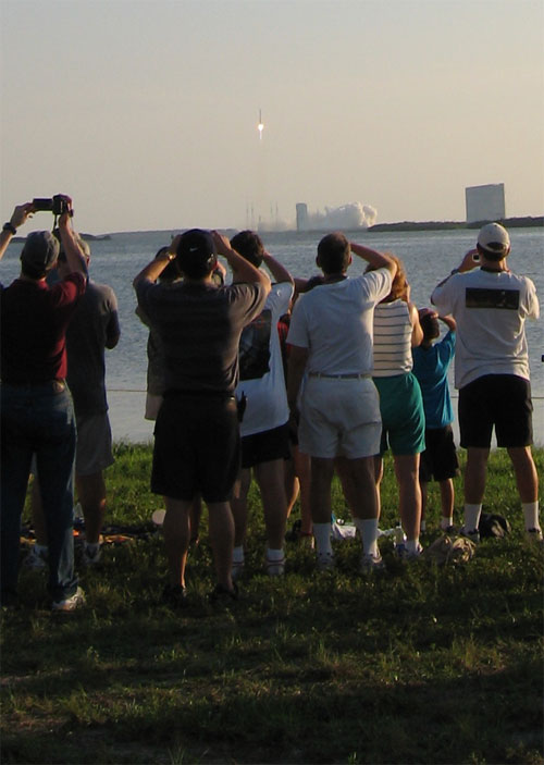 In this image, a handful of the many guests watching the Mars Reconnaissance Orbiter launch stand near the shore of the Banana River that separates the launch pad at Cape Canaveral Air Force Station from Kennedy Space Center.  Most people are holding up cameras to capture the moment.  In the center of the image is the rocket lifting from its pad.  From this distance (a few miles), the rocket looks like a thin line in the sky with orange fire spitting from the bottom.  Smoke billows on the ground around the launch pad.