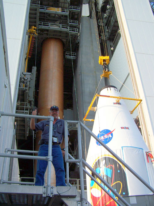 JPL's launch vehicle manager Arden Acord, a man in his mid-forties with gray hair and gray beard and goatee, wears a dark colored hat and huge smile as he stands next to the large, brightly-decorated, protective fairing for the Mars Reconnaissance Orbiter.  The fairing bears the red, white and blue circular NASA logo as well as the colorful depiction of the spacecraft at work around Mars.  The fairing is attached to a rig that will lift it up to be attached to the top of the Atlas V rocket that will launch it to Mars.  Behind Acord and the fairing is the Atlas V rocket in its extremely tall Vertical Integration Facility.