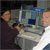This photo shows Ruth Fragoso and Glen Havens turning from their shared computer screens that are spilling out lines and lines of spacecraft data to give a thumbs up and a smile to the camera.  Ruth is tan with shoulder-length, shiny black hair, and wears a dark plum-colored suit and a genuine smile with a cute dimple.  Glen has short blond hair, blue eyes, and a broad smile.  He wears a white dress shirt and a silver tie.  A blue sign with white letters, spelling MOSE, sits on top of their computer screens.