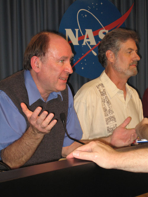 In this image, two Caucasian men are shown, seated in front of a bluish-gray drapery backdrop. The circular NASA 'meatball' logo is between them and the backdrop.  On the left is the Mars Reconnaissance Orbiter project scientist Rich Zurek, a balding man with brown hair and green eyes. He is wearing a blue short-sleeved shirt under a gray sweater vest.  His hands are raised as if he's explaining something to the reporter (not pictured).  In the right of the image is the principal investigator for the HiRISE camera aboard the Mars Reconnaissance Orbiter, Alfred McEwen.  He is a middle-aged man with a graying beard.  He is wearing a cream-colored 'Hawaiian' shirt.
