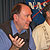 In this image, two Caucasian men are shown, seated in front of a bluish-gray drapery backdrop. The circular NASA 'meatball' logo is between them and the backdrop.  On the left is the Mars Reconnaissance Orbiter project scientist Rich Zurek, a balding man with brown hair and green eyes. He is wearing a blue short-sleeved shirt under a gray sweater vest.  His hands are raised as if he's explaining something to the reporter (not pictured).  In the right of the image is the principal investigator for the HiRISE camera aboard the Mars Reconnaissance Orbiter, Alfred McEwen.  He is a middle-aged man with a graying beard.  He is wearing a cream-colored 'Hawaiian' shirt.