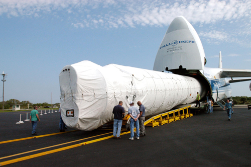 This image shows the Atlas V rocket booster being delivered to Cape Canaveral. Under blue skies streaked with thin clouds, technicians supervise the unloading of the large Atlas V rocket booster from a Russian cargo plane.  The booster is covered in a protective white cloth.  Looking like a great white shark with its mouth wide open, the enormous cargo plane's nose cone is lifted to allow the rocket to be rolled out onto the asphalt 'Skid Strip' at Cape Canaveral.