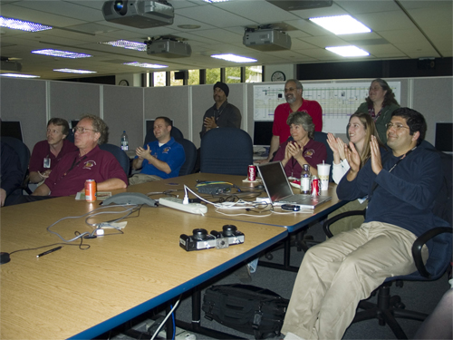 In this image, a group of nine people sit at a large, rectangular table, viewing a monitor that is showing NASA television.  The men and women are clapping.