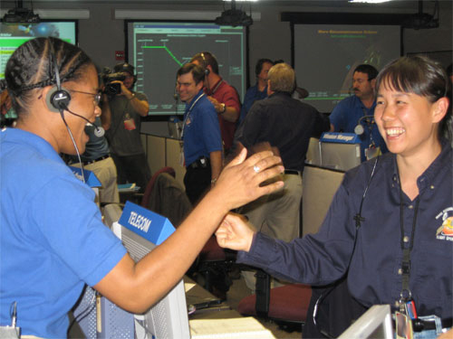 In this image, Mars Reconnaissance Orbiter System Engineer Tracy Drain (an African-American woman) shakes the hand of her co-worker, Telecommunications Engineer Ramona Tung (an Asian-American woman).  Tracy wears a bright blue short-sleeved shirt and Tung wears a dark blue long-sleeved shirt with the MRO mission logo on it.  They are in the mission control area at JPL - a room with rows of computer consoles and large screens that display mission data.  They are surrounded by dozens of other team members celebrating.