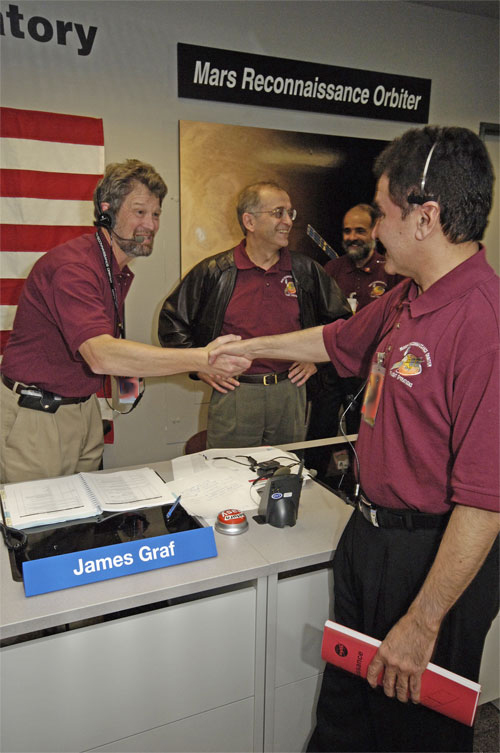 In this image, Graf, a middle-aged Caucasian man with brown hair and a goatee firmly shakes the hand of Naderi, a middle-aged Persian-American man with dark brown hair.  Dr. Charles Elachi, Director of the Jet Propulsion Laboratory (a middle-aged Lebanese-American man) is in the background.  They are all wearing short-sleeved, maroon polo shirts with the mission logo on them.