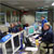 In this image, a group of seven engineers sit at their computer consoles, which are set up in 4 parallel rows.  Each station has a blue triangular sign atop it that identifies the job function of the person siting there.  For example: telecommunication, attitude control, navigation, etc.  In the background are two large screens that display mission data.  The engineers are all wearing headsets that they use to communicate with one another.
