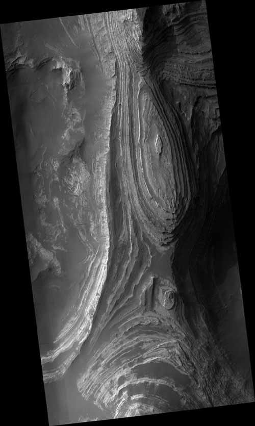 This black-and-white orbital image shows elongate, teardrop-shaped layers of rock stacked atop each other and viewed from above. To the left and right, the layers drop off as sharp cliffs to sandier surfaces below.