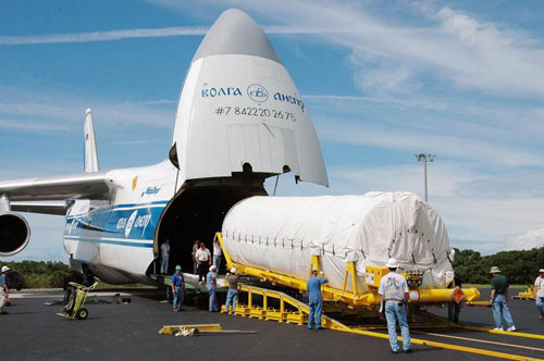 Looking like the mouth of an enormous great white shark, a white and blue Russian cargo plane is opened to reveal the large Centaur upper stage engine that will blast the Mars Reconnaissance Orbiter to Mars.  The large cylindrical engine is covered in a protective white tarp and is being carefully marshaled off the plane and down a bright yellow ramp by a team of technicians.  The bright blue sky, streamed with wispy clouds is in the background along with a line of brilliant green trees.