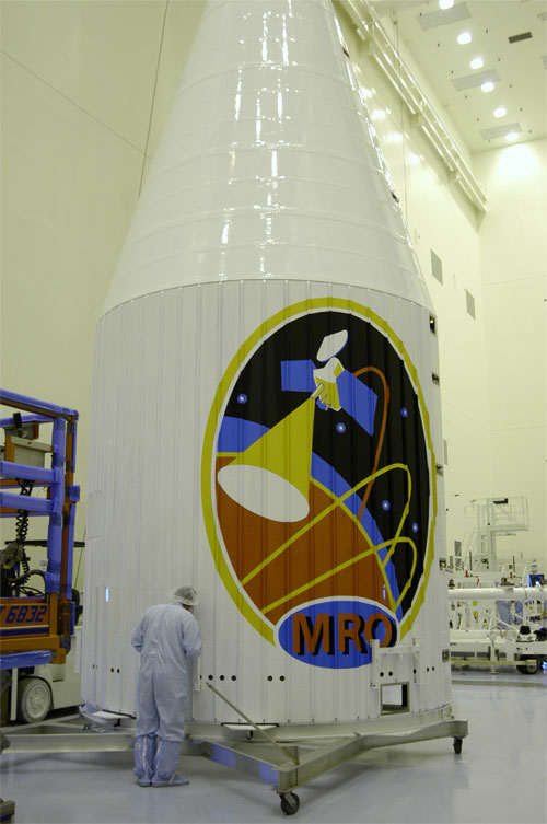 This image features the large nose cone that will protect the Mars Reconnaissance Spacecraft before and during launch.  The white structure's larger, lower cylinder is capped by a cone-shaped piece.  The colorful Mars Reconnaissance logo adorns the side.  The logo background is black and dotted with simulated stars.  The cartoon spacecraft is 'conducting science' and is featured with brightly colored swirls that simulate the spacecraft's orbit around the planet Mars.  Within the logo, the swath of the HiRISE camera is simulated and that swath echoes the cone shape of the fairing itself.  A person dressed in light blue cleanroom attire (called a 'bunny suit') stands in front of the fairing, which is nearly four times his or her height.