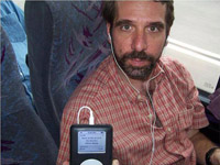 This picture of Jeff Plaut was taken inside a bus.  Jeff is in his late-thirties, has brown hair and a beard, and wears a red, short-sleeved, button-up shirt.  He's looking directly into the camera, slightly smiling, and holding up a portable music player to show the digital display, which says, 'Back in the U.S.S.R.  The Beatles.  White Album.'  Small white headphones are in Jeff's ears and attach by a chord to the music player.