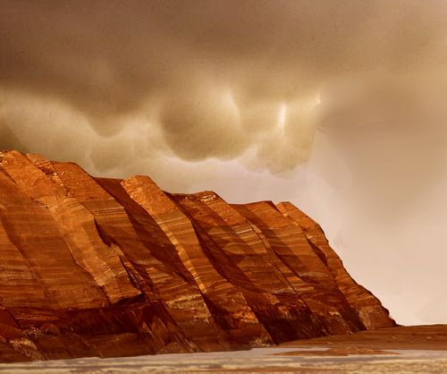 This image shows layered, red cliffs rising up from a flat, brown surface. The cliff surface slants from right to left, with the nearest cliffs on the right-hand side of the image and those farther away toward the left. Pinkish-tan clouds hover above the crests of the cliffs.