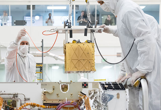 In a bright white cleanroom facility, technicians carefully lower a gold-colored cube – the MOXIE instrument – into the body of the Mars rover Perseverance.